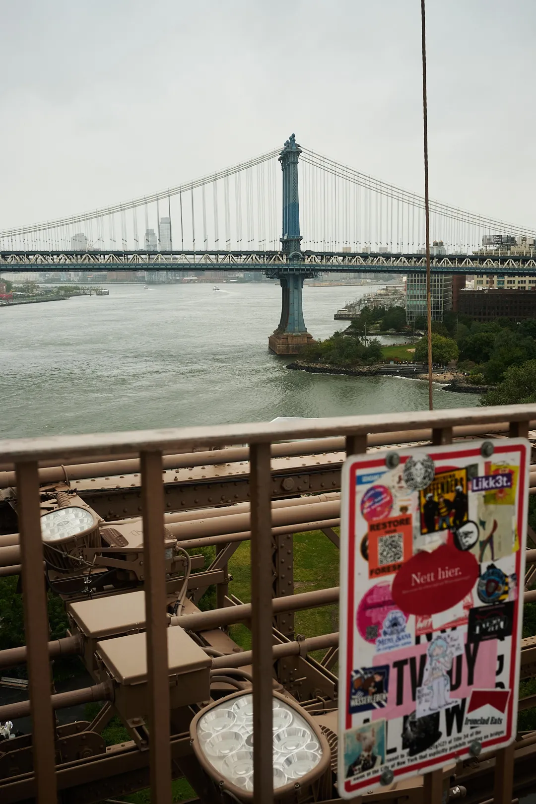 A sign on the Brooklyn Bridge covered in stickers, the Manhattan Bridge behind